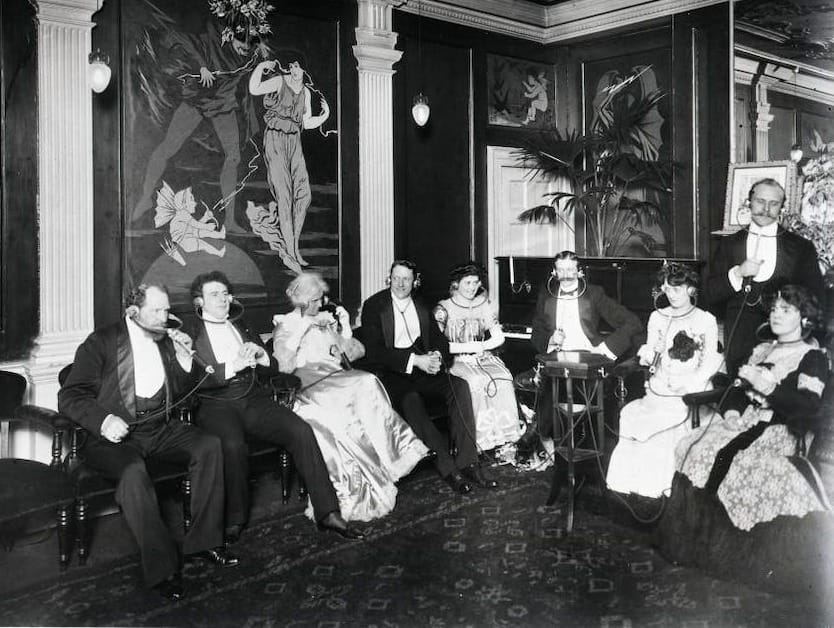 A group of well-dressed men and women from the period listening to infotainment through an Electrophone.