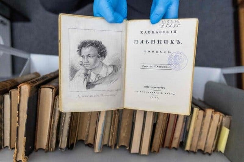 A pair of hands in blue rubber gloves holds up a fake copy of a firsts edition of a Pushkin book. The book is held open, showing Cyrillic writing and a black and white image of a young man. 